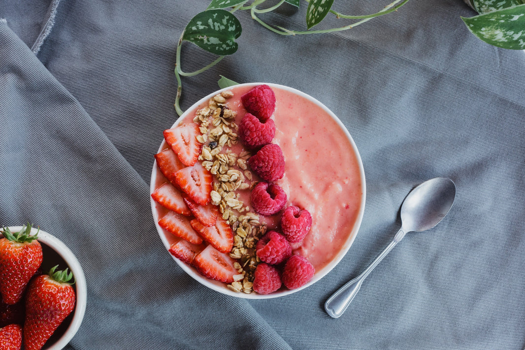 Pink smoothie bowl topped with fresh strawberries, raspberries, and granola on gray linen with additional berries